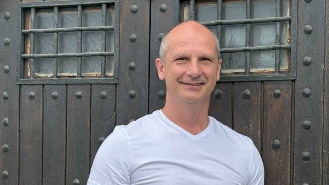 A business scaling coach with a shaved head, wearing a white t-shirt, stands in front of a large wooden door with metal bars and rivets.