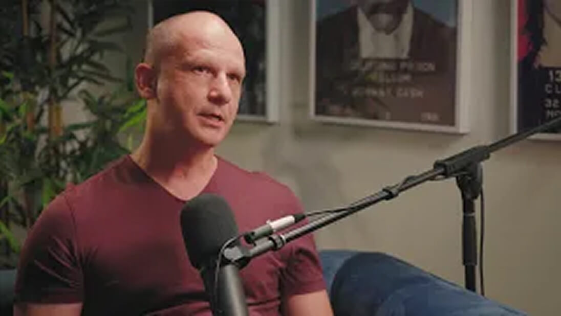 A bald man in a maroon shirt sits on a blue couch, speaking into a microphone during a podcast or interview as a business scaling coach, with framed posters on the wall behind him.